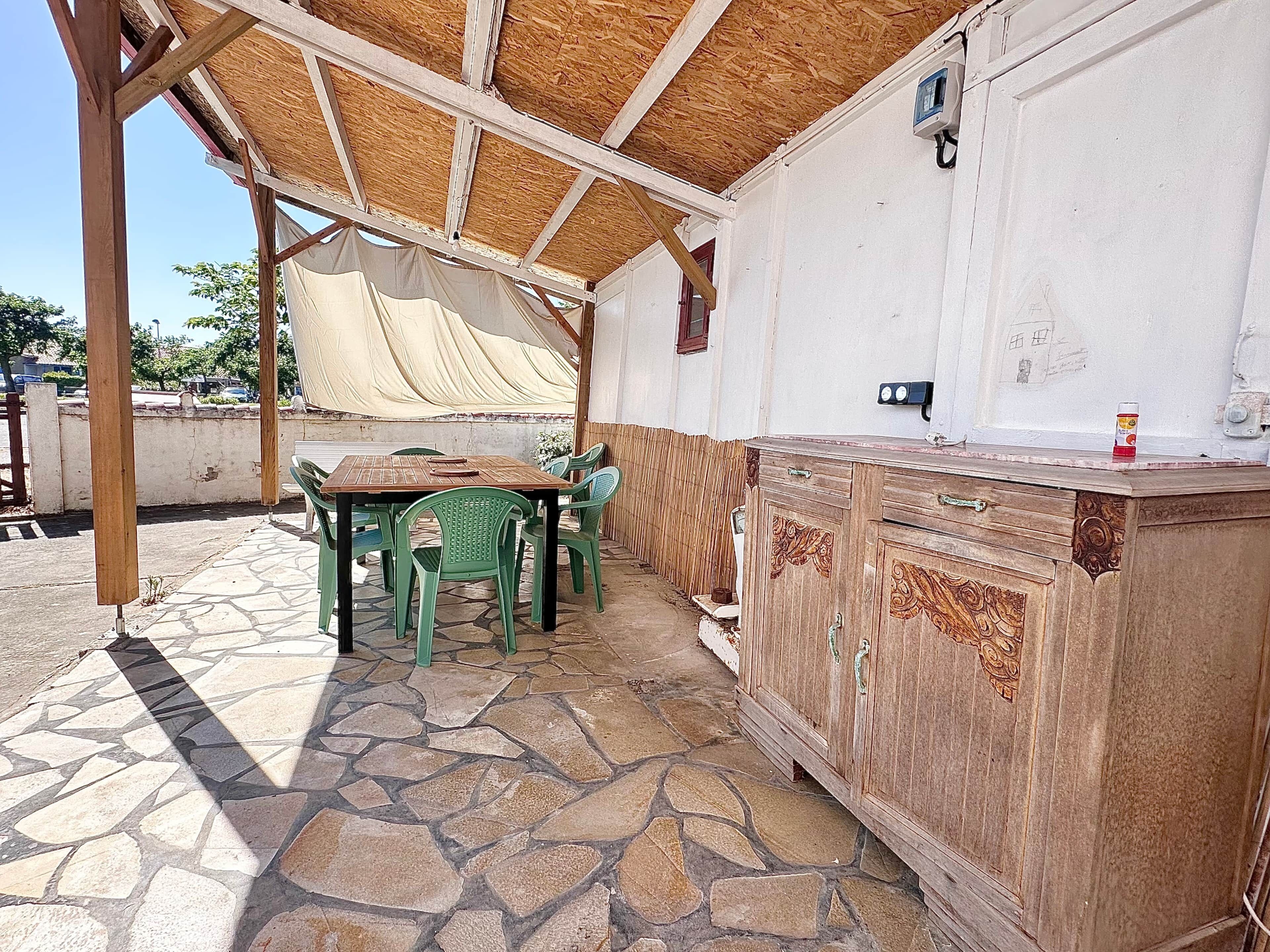 An outdoor patio with a stone floor, a wooden table, green plastic chairs, and a wooden cabinet.