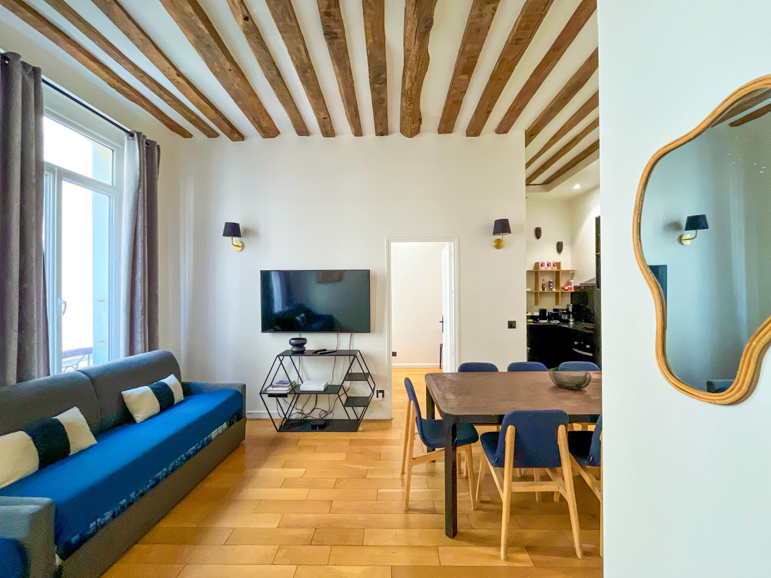 Una sala de estar con un sofá, un televisor sobre un mueble bajo, una mesa de comedor con sillas, piso de madera y un techo con vigas de madera.