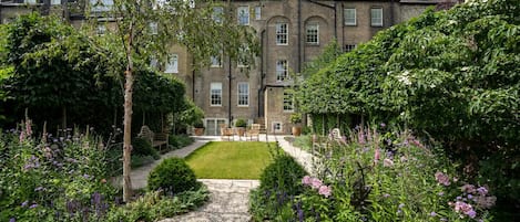 A traditional brick building with a symmetrical facade, a central lawn, and a variety of flowering plants.