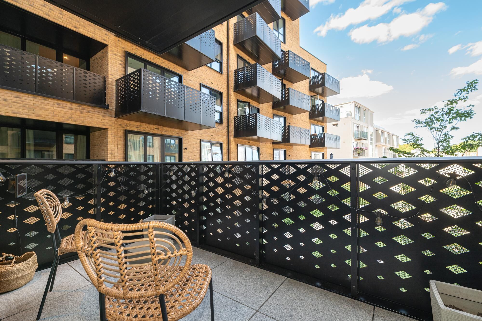 Un balcón con una silla de mimbre, un panel perforado negro y vistas de un edificio moderno con balcones.