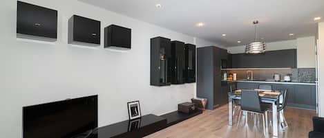 Uma sala de estar moderna com televisão de tela plana, lareira, área de jantar com mesa e cadeiras, e cozinha ao fundo.
