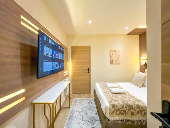 Habitación de hotel con un televisor de pantalla plana grande montado en la pared, una puerta de madera, una cama con cubrecama estampado, una mesita de noche pequeña y una alfombra con diseño.