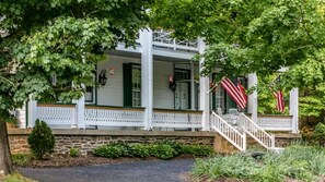 A covered porch with a stone foundation, white railings, and two American flags. - Exclusive Whole-Inn Lodging: The Ultimate Shenandoah Valley Group Retreat at The Buckhorn (Churchville)