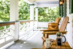 Ein Holztisch mit zwei Kaffeetassen und einer Kaffeemaschine auf einer Veranda mit Geländer, mit Blick auf einen Garten.