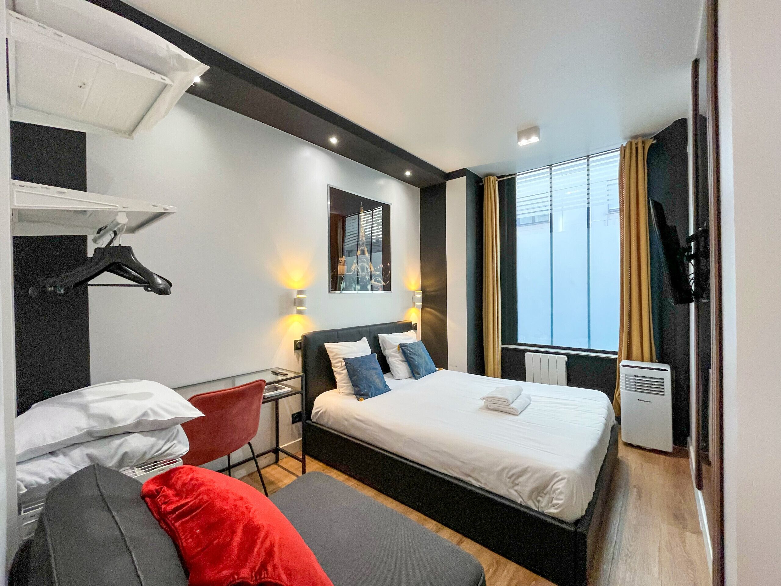 A modern hotel room with a bed, a desk, and a chair. There is a window with curtains, a wall-mounted TV, and a heater.
