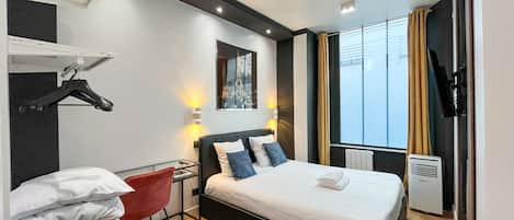 A modern hotel room with a bed, a desk, and a chair. There is a window with curtains, a wall-mounted TV, and a heater.