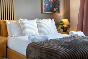 A neatly made bed with white bedding and striped pillows, a bedside lamp, a potted plant, and a painting on the wall. - Modern Riverside Flat in City Centre, Free Parking - Pass the Keys (Cambridge)