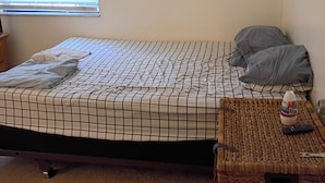 A single bed with a checkered bedspread, a pillow, a remote control, and a water bottle on a woven tray.