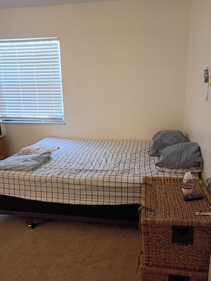 A single bed with a checkered bedspread, a pillow, a remote control, and a water bottle on a woven tray.