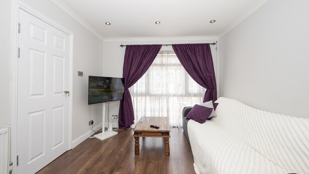 Ein Schlafzimmer mit weißer Tür, einem Fernseher, einem Holztisch, einem weißen Sofa und violetten Vorhängen.