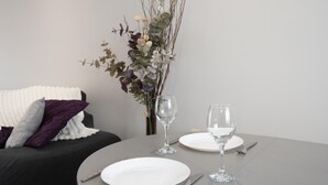 Ein Esstisch mit zwei weißen Tellern, zwei Weingläsern und Besteck. Auf dem Tisch steht eine Vase mit getrockneten Ästen und Blumen.