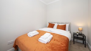 Ein ordentlich bezogenes Bett mit weißer und orangefarbener Bettwäsche, eine Nachttischlampe und eine kleine Pflanze auf einem Nachttisch.