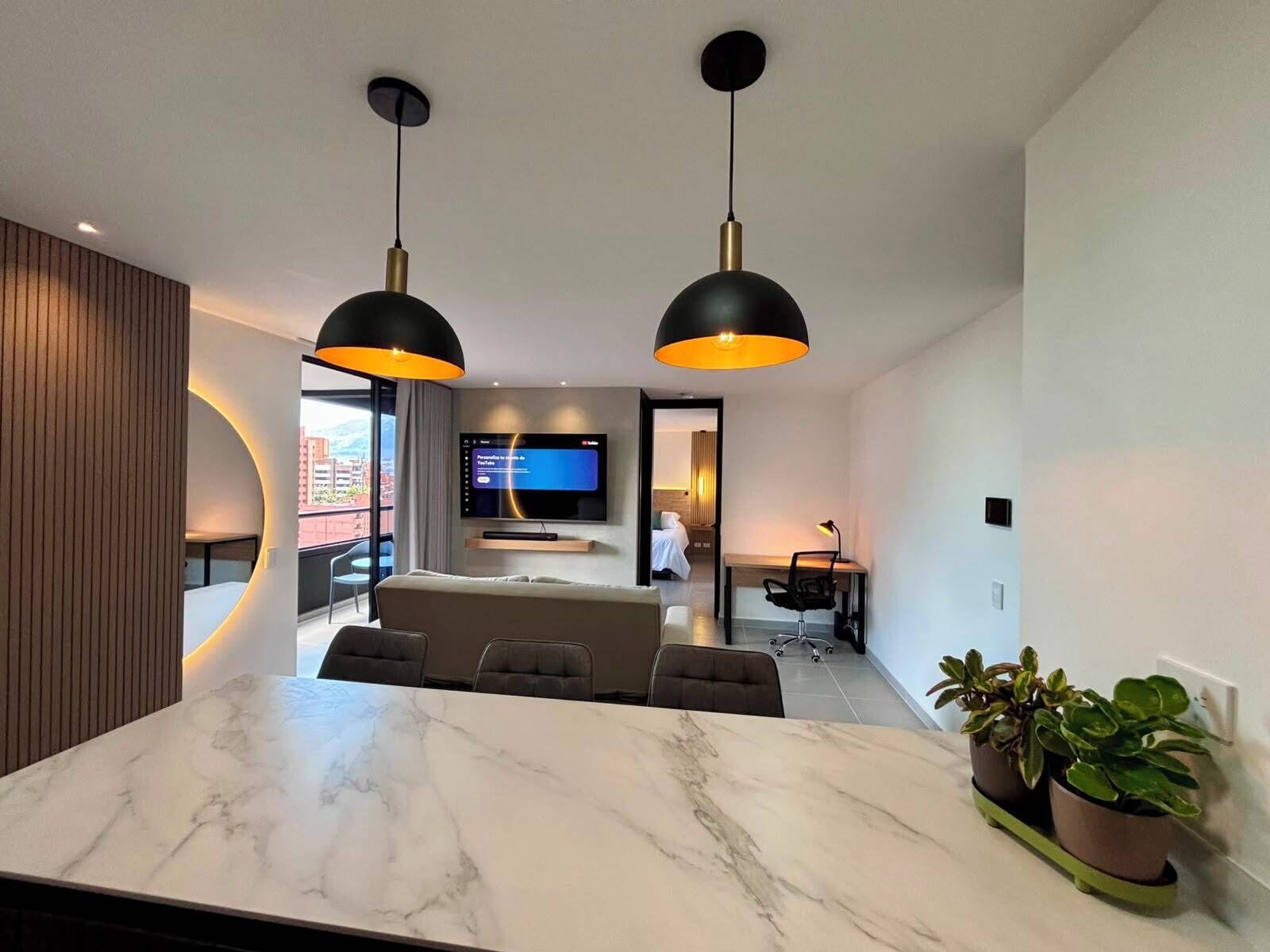 Une chambre d’hôtel moderne avec une table à manger en marbre, deux lampes suspendues, un téléviseur à écran plat, un canapé et un bureau avec une chaise.