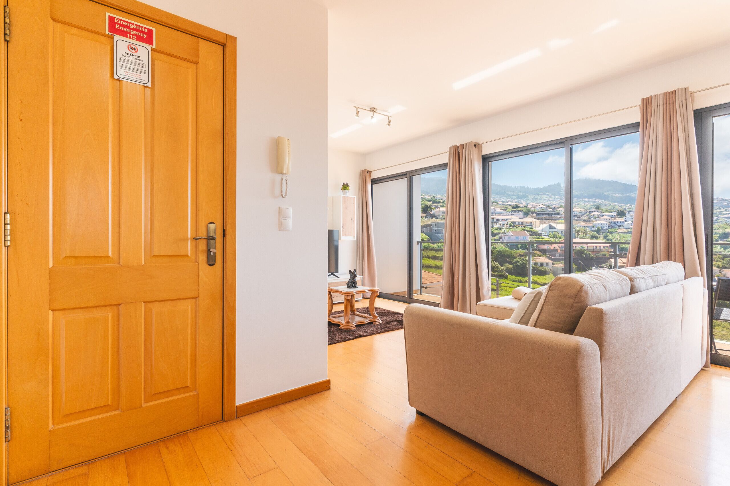 A hotel room with a wooden door, a sofa, a small table, a plant, and a view of a town through large windows.