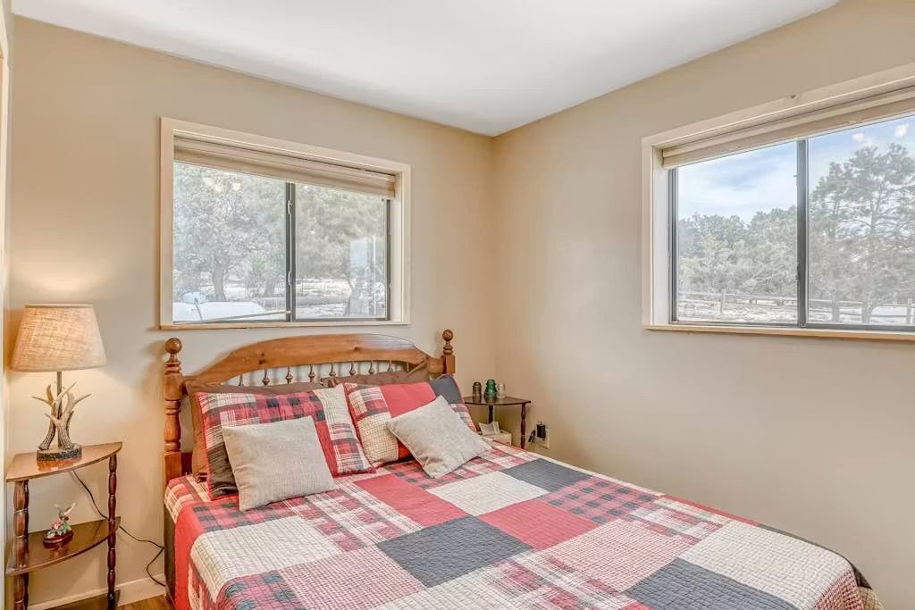 A bedroom with a wooden bed, a nightstand, a lamp, and two windows with a view of trees.