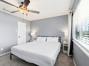 A bedroom with a bed, a ceiling fan, a nightstand, a lamp, and a window with blinds.