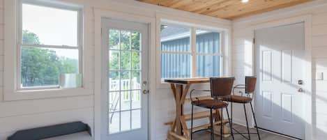 A room with a wooden ceiling, a bench, a table with chairs, and a view of a blue fence through a window.