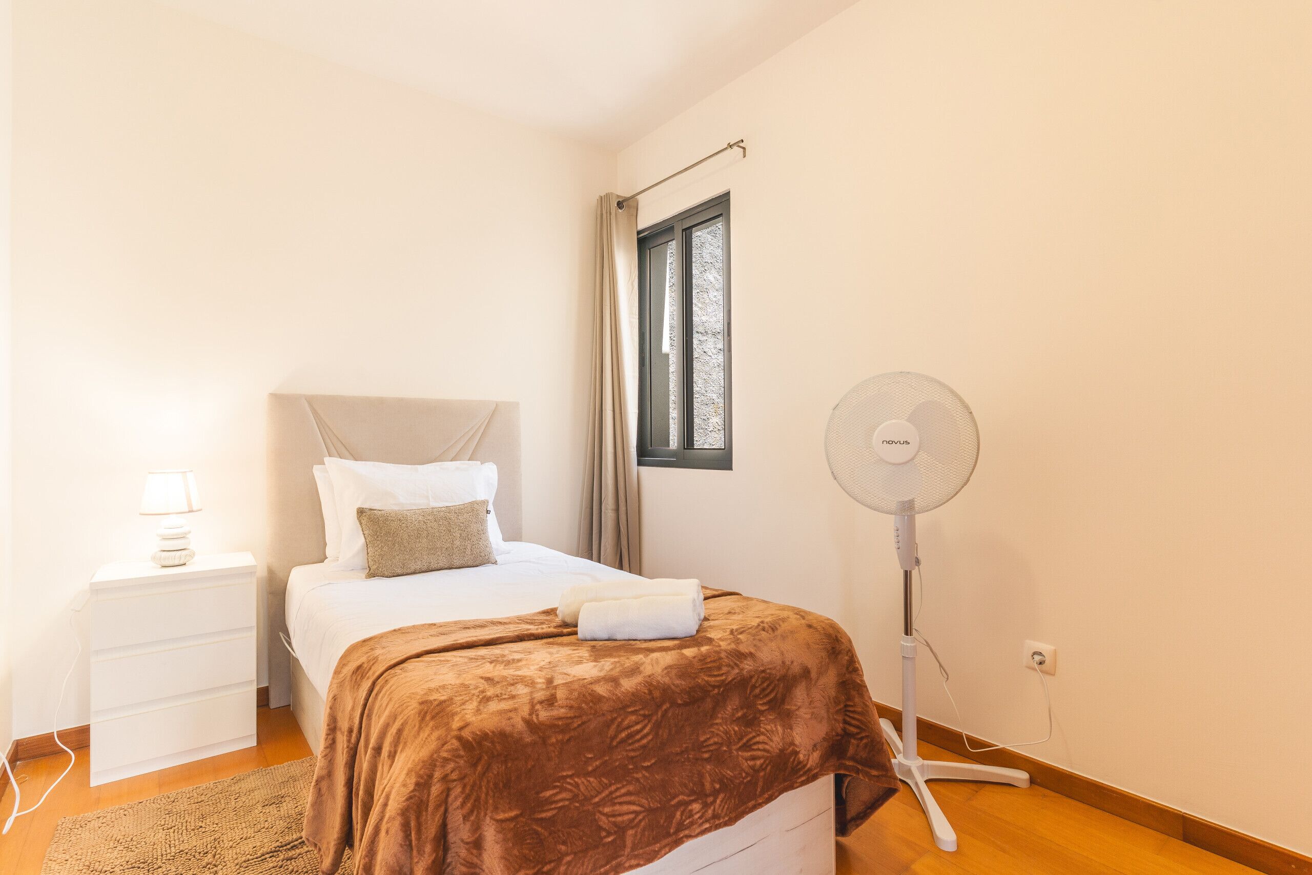 A bedroom with a bed, bedside table, lamp, and a standing fan.