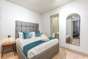 A modern hotel room with a large bed, a bedside table, a mirror, and a bathroom visible through an open door. - One Rosebank | Sophisticated City Living (Johannesburg)