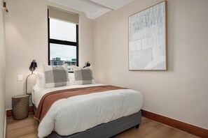 A bedroom with a bed, a window, a bedside lamp, a trash bin, and a large abstract painting on the wall. - Charlie and the Chairman | 2 Bedroom City Escape (Cape Town)