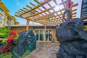 Une pergola en bois marque l’entrée d’un restaurant et d’un bar à l’ambiance tropicale, avec de grands rochers et un sol carrelé.
