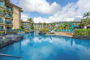 A resort pool area with multiple lounge chairs and umbrellas, surrounded by a building with balconies and palm trees. - New Listing- Waipouli Ocean front, Pool, AC, GYM, Waterslide! (Kapaa)