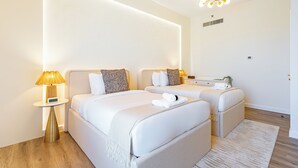 A modern hotel room with two beds, a bedside table with a lamp, a small bedside table with a clock, and a ceiling light fixture. - Upgraded Modern 3BR+Kids | JBR | Sea and Ain View (Dubai)