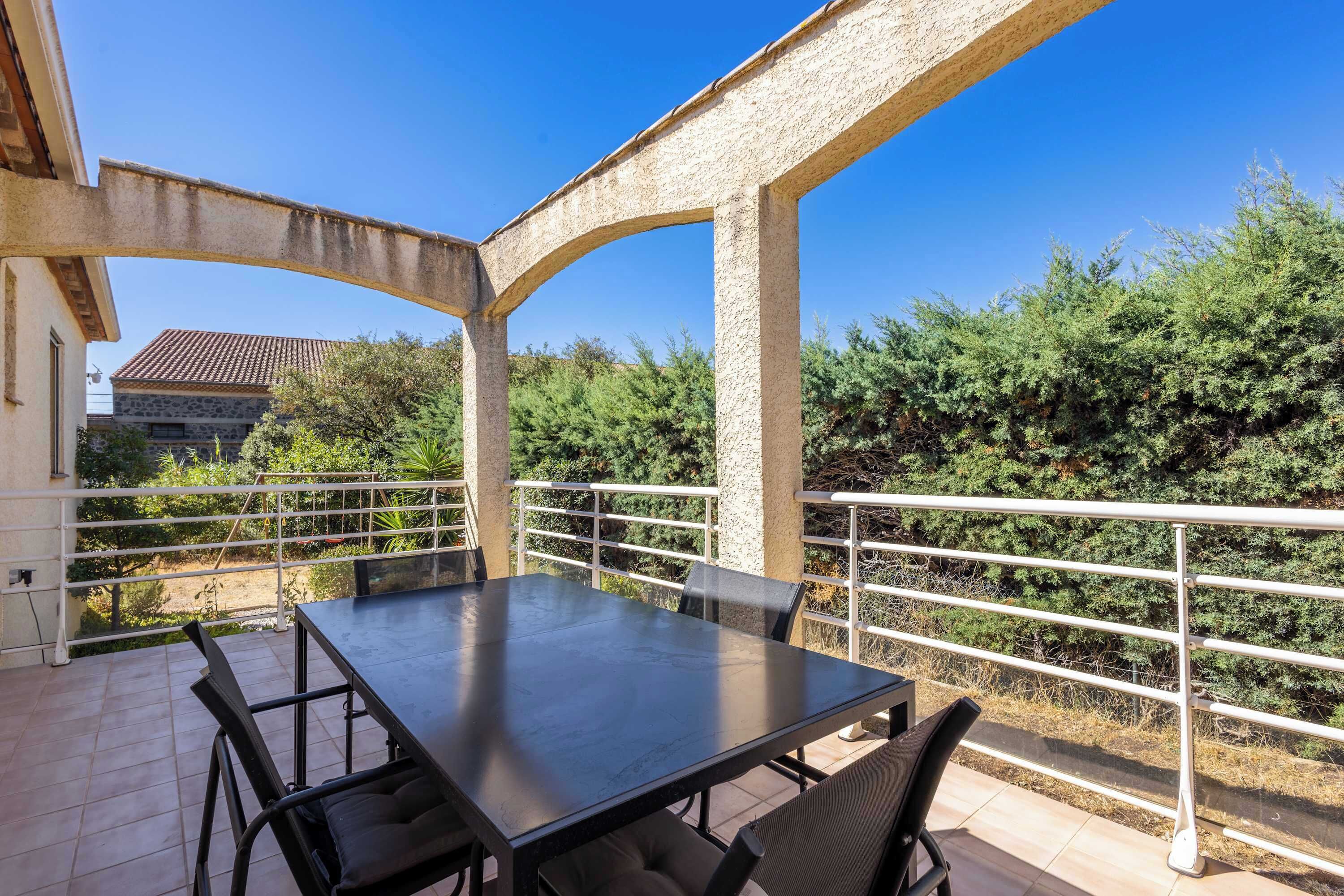 A terrace with a table and chairs, surrounded by a white railing, and a view of greenery and a building in the background.