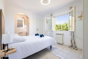 Ein Schlafzimmer mit einem Bett, einem Nachttisch mit Lampe, einem Fenster mit Blick auf Grünflächen und einer Heizung.