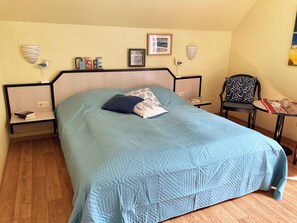 Ein ordentlich bezogenes Bett mit einer blauen Decke, zwei Nachttische mit Lampen, ein Sessel und ein kleiner Tisch mit einem Buch und einer Taschentuchbox.