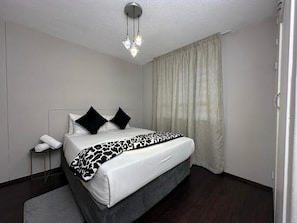 Habitación de hotel con cama, mesita de noche, lámpara y ventana con cortinas.