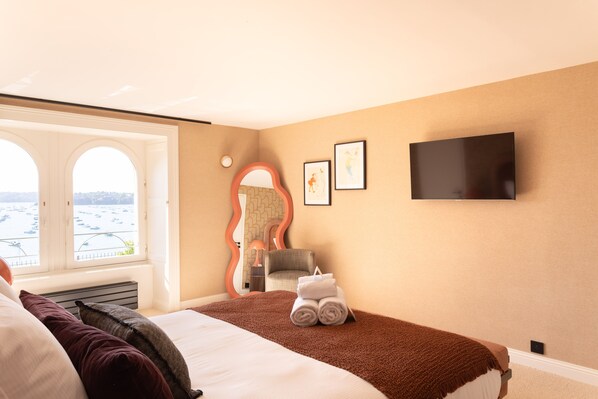 Habitación de hotel con cama, televisión, espejo y vista al mar por una ventana.