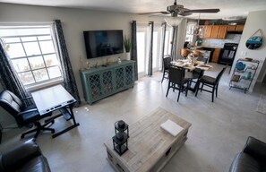 A modern living room with a flat-screen TV, a dining area with a table and chairs, a kitchen with wooden cabinets, and a ceiling fan. - Luxury Waterfront Condo in Coquina Key w/ Breathtaking Views - gated community. (St. Petersburg)