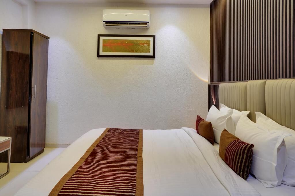 A hotel room with a bed, a wardrobe, an air conditioning unit, a framed picture, and a headboard with vertical slats.