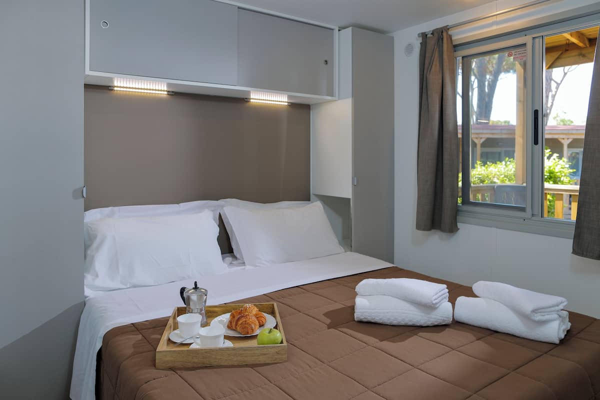 Una cama bien hecha con sábanas blancas, una bandeja con un croissant, una taza y una manzana, y una ventana con cortinas.