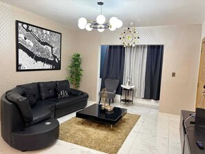 A modern living room with a black sofa, a coffee table, a chandelier, a large abstract wall art, a potted plant, and a view of the outdoors through a window with curtains. - Apartment in Arismar (Santo Domingo Este)