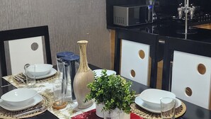Un comedor con una mesa larga dispuesta para cuatro personas, rodeada de sillas. Se observa una cocina al fondo con un microondas y una estufa.