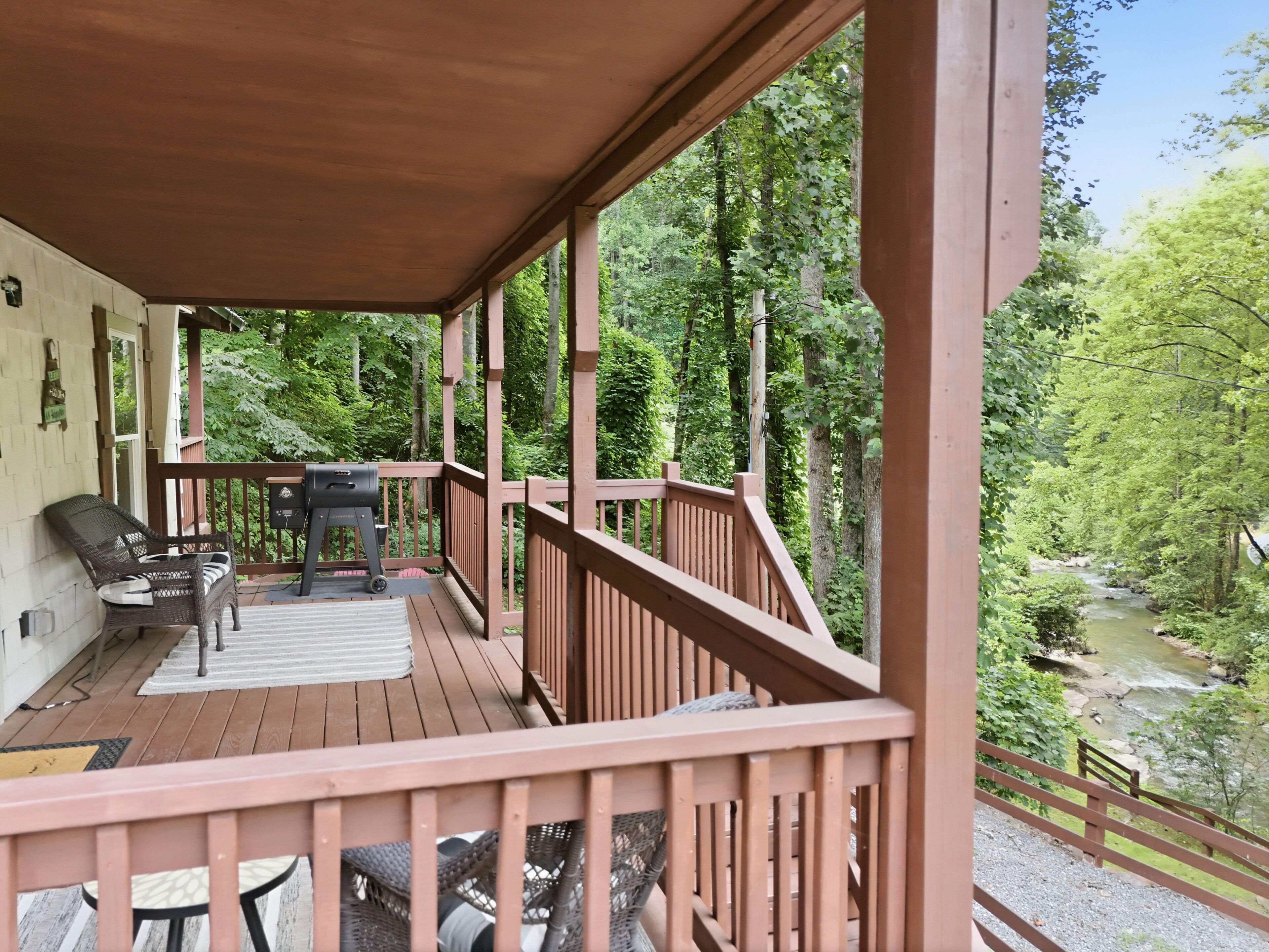 Une terrasse en bois, dotée de mobilier d’extérieur, d’un barbecue et offrant une vue sur une zone boisée traversée par un ruisseau.