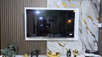 A modern living room with a flat-screen TV mounted on a wall with a marble pattern. Below the TV, there is a white entertainment unit with cabinets and a shelf holding decorative items.