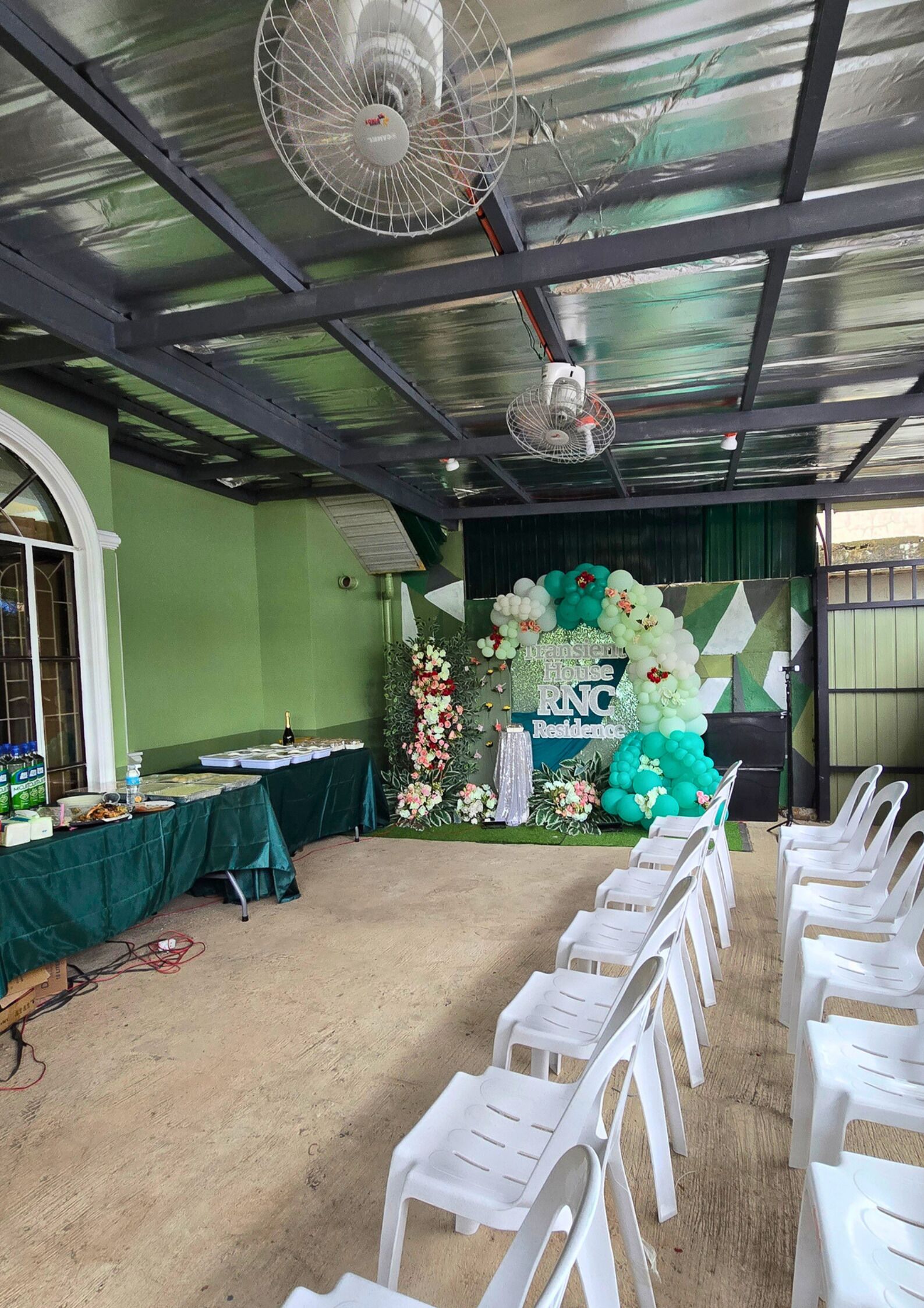 Un espace extérieur abrité, aménagé pour les événements, avec une scène décorée de fleurs et de ballons, une table garnie de nourriture et de boissons, et des rangées de chaises en plastique blanches.