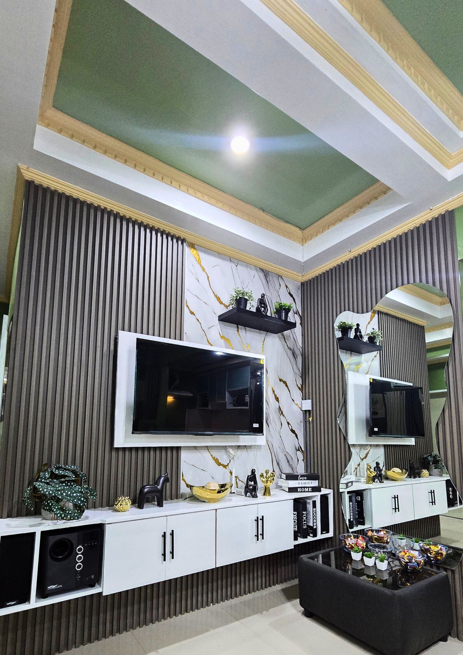Un salon moderne doté d’un grand téléviseur à écran plat fixé sur un mur en marbre, d’un canapé noir et d’un meuble de rangement blanc contenant des livres et des objets décoratifs.