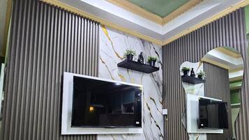 Un salon moderne doté d’un grand téléviseur à écran plat fixé sur un mur en marbre, d’un canapé noir et d’un meuble de rangement blanc contenant des livres et des objets décoratifs.