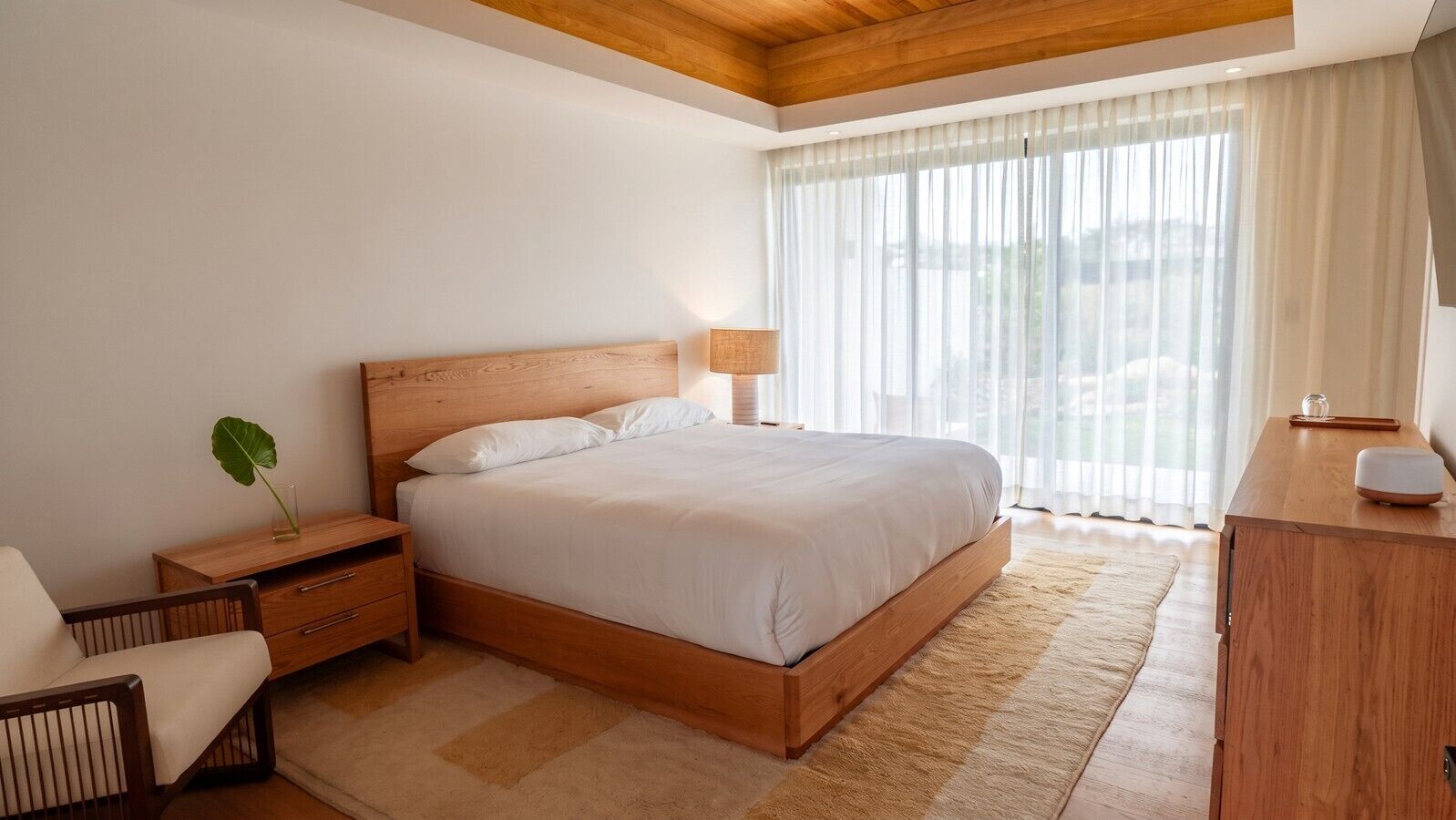Een slaapkamer met een houten bed, nachtkastjes, een bureautje en een stoel, en een groot raam met dunne gordijnen.