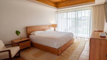 A bedroom with a wooden bed, bedside tables, a nightstand, a chair, a desk, and a large window with sheer curtains.