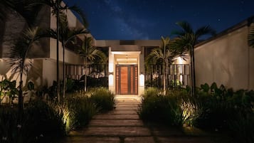 A nighttime view of a building entrance with a wooden pathway leading to a door, flanked by palm trees and illuminated by wall-mounted lights.