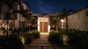 Vista nocturna de la entrada de un edificio con un camino de madera que conduce a una puerta, flanqueada por palmeras e iluminada por luces empotradas.