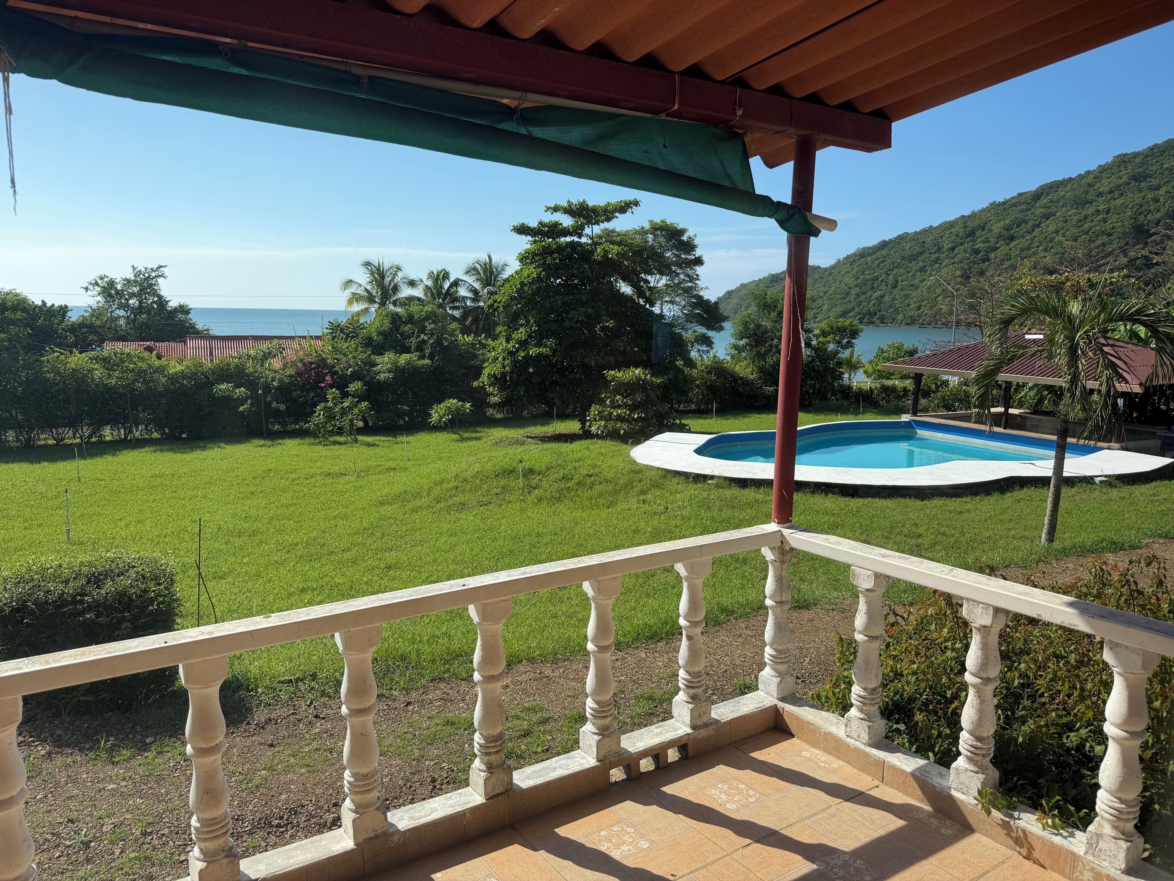 Ein Balkon mit Blick auf einen Swimmingpool, üppiges Grün und ein entferntes Gewässer.