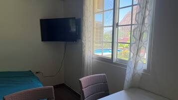 Ein Zimmer mit einem Bett, einem Sessel, einem Tisch, einem an der Wand befestigten Fernseher und einem Fenster mit Blick auf ein Gebäude und einen Pool.