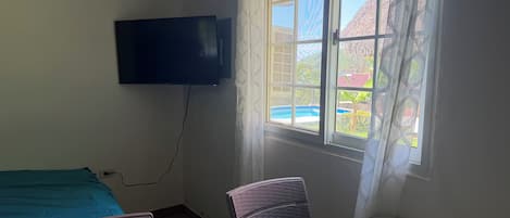 A room with a bed, a chair, a table, a TV mounted on the wall, and a window with a view of a building and a pool.