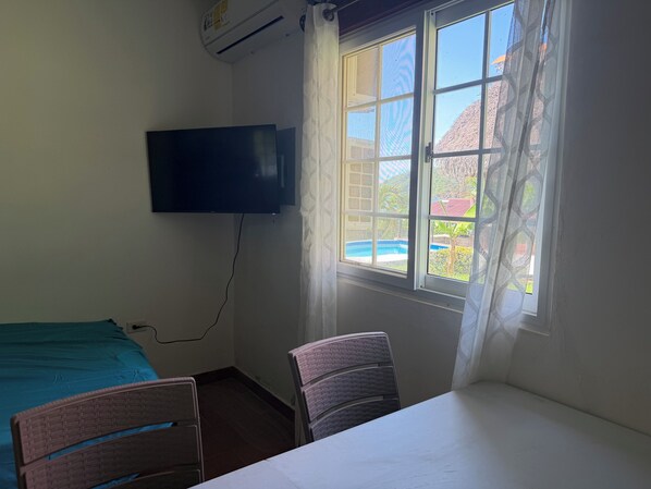 A room with a bed, a chair, a table, a TV mounted on the wall, and a window with a view of a building and a pool.